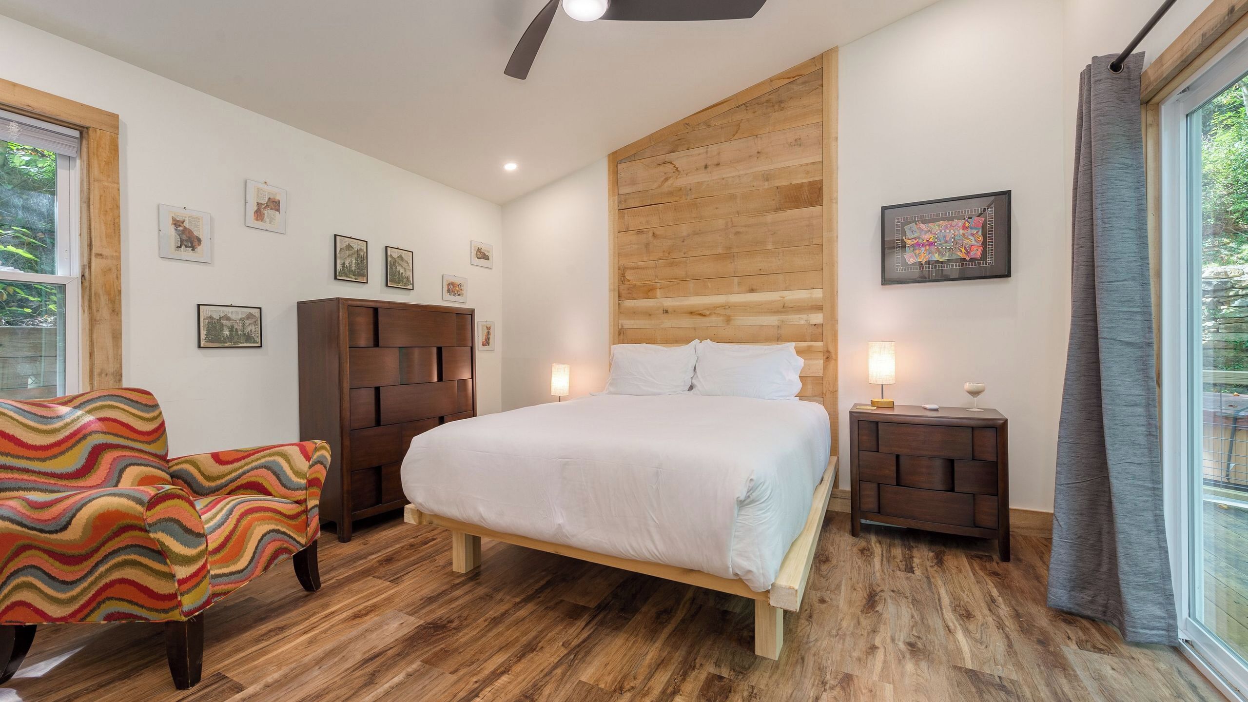 A bedroom with a bed, a wooden headboard, a nightstand with a lamp, a colorful chair, and framed pictures on the wall.