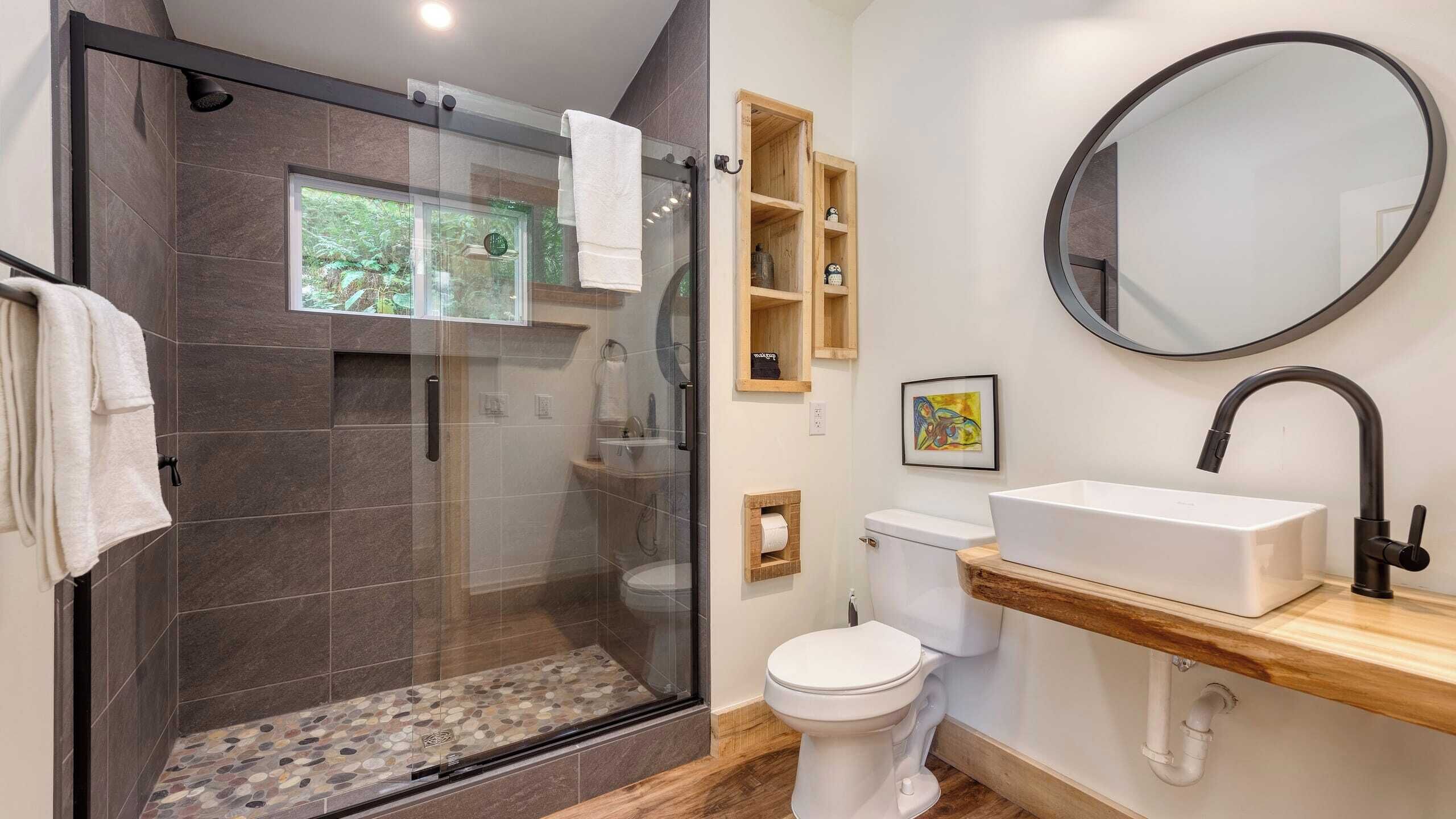 A modern bathroom with a glass-enclosed shower, a white toilet, a wooden vanity with a sink, and a round mirror.