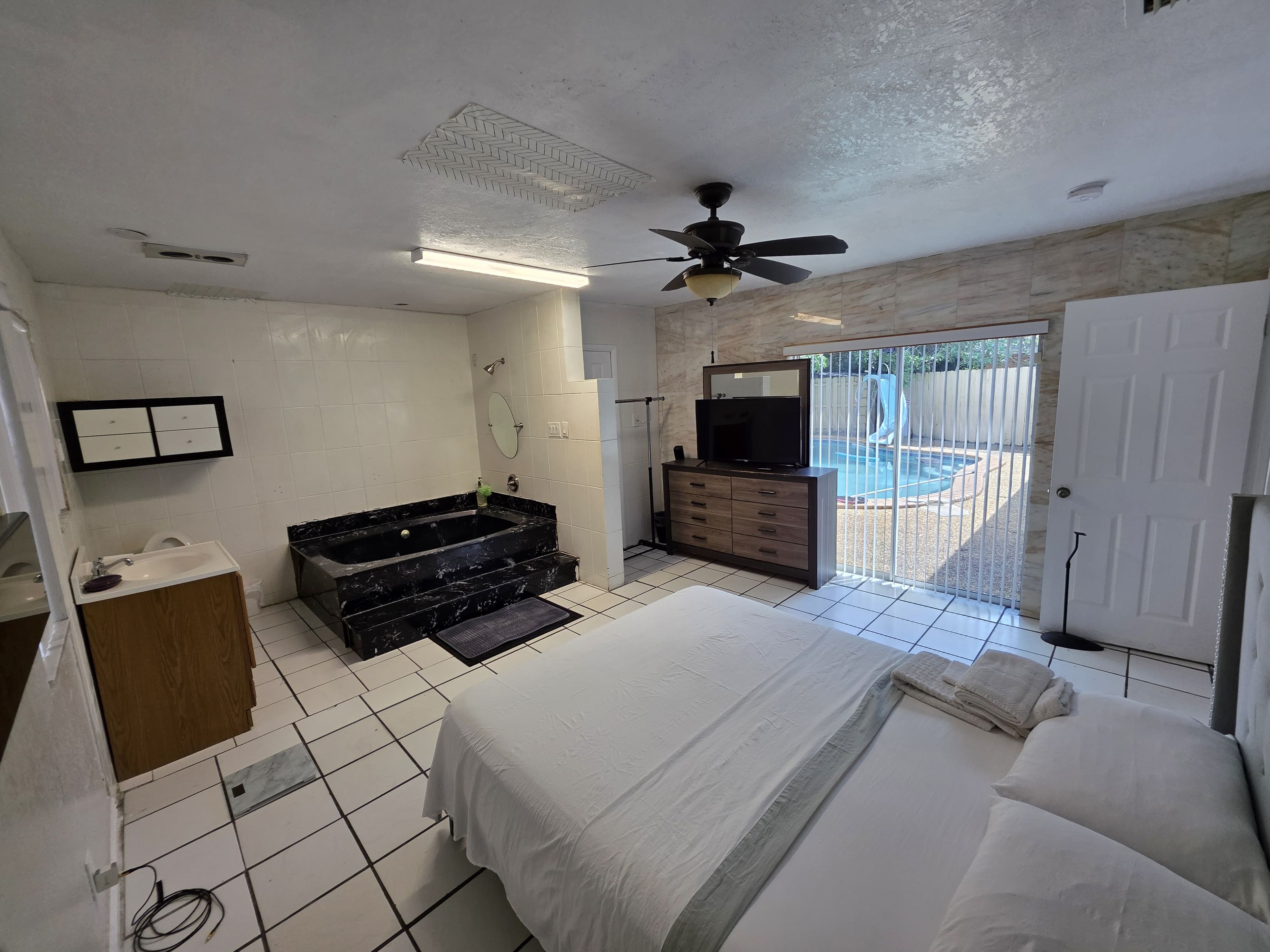 A bedroom with a bed, a vanity, a bathtub, a ceiling fan, and a view of an outdoor swimming pool.