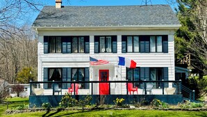A two-story house with a red door, white siding, and a balcony with chairs.