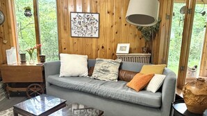 A cozy room with a grey sofa, a wooden coffee table, and a large window with a view of greenery.