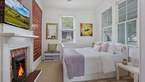 A bedroom with a fireplace, a bed, a nightstand, a window with blinds, and a painting on the wall. - Redstick Cottage (Baton Rouge)