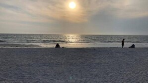 En strand med solnedgang over vandet, mennesker sidder og går langs kysten.