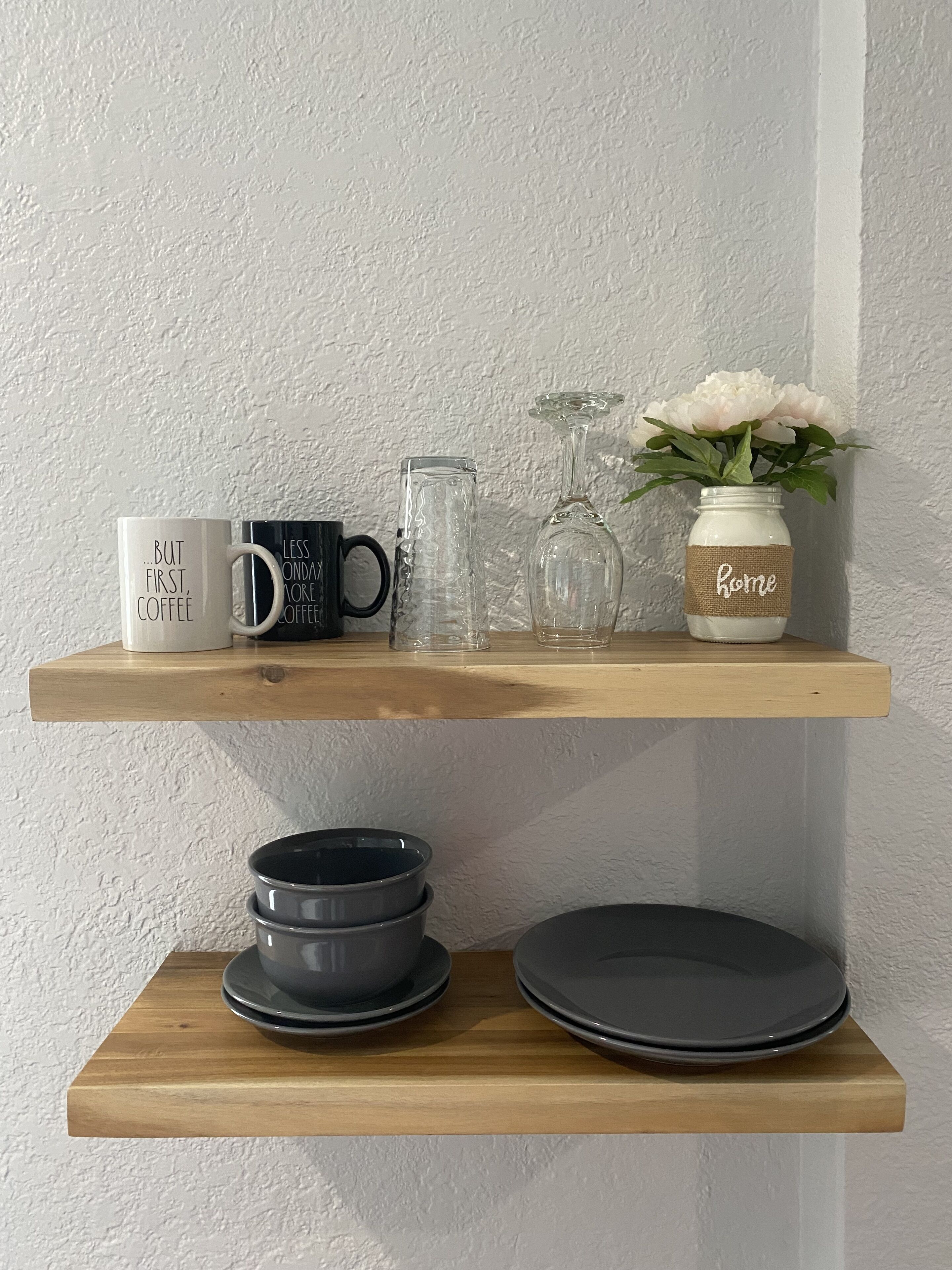A wooden shelf with a white mug, a black mug, a glass, and a vase with flowers. Below, there are stacked plates and bowls.
