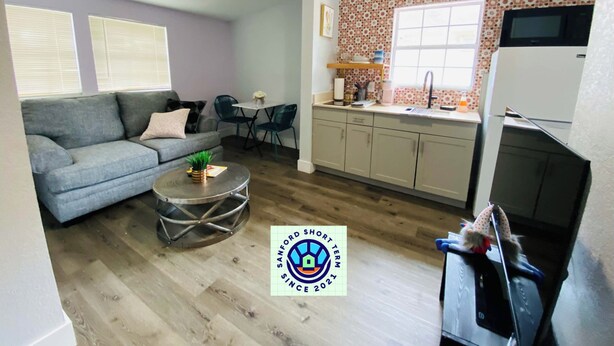 A compact living space with a grey sofa, a small round coffee table, a kitchen area with a tiled backsplash, and a refrigerator. - The Cherry Blossom (Sanford)