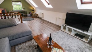 Un salon chaleureux avec un canapé gris, une table basse en bois sur laquelle reposent un livre et des lunettes, un téléviseur à écran plat et une lucarne.