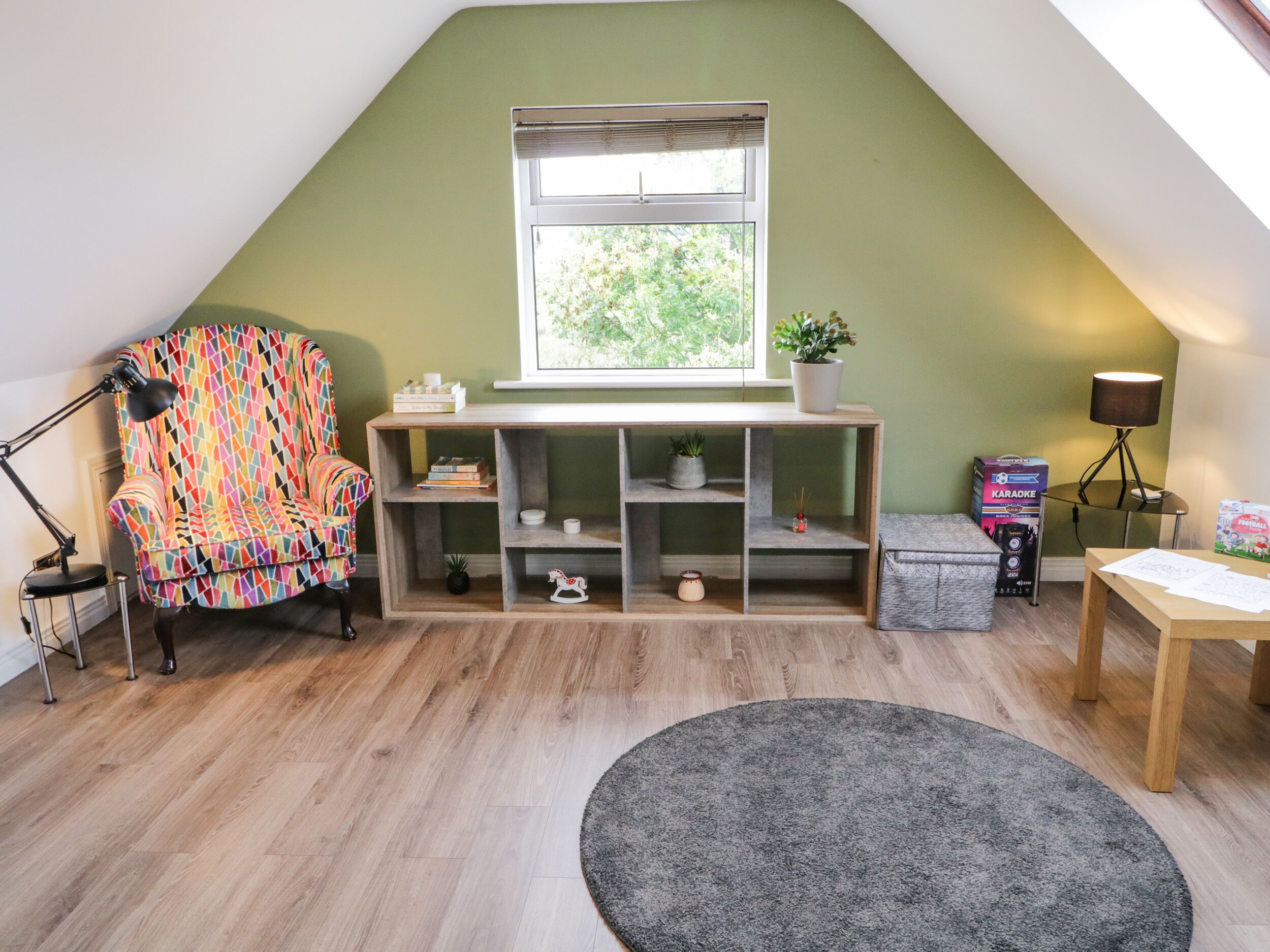 Une pièce chaleureuse avec un plafond en pente, un fauteuil coloré, une console en bois, un bureau avec des papiers, une lampe et une fenêtre donnant sur un paysage verdoyant.
