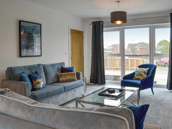 A modern living room with a grey sofa, a blue armchair, a glass coffee table, and a large window with a view of a residential area.