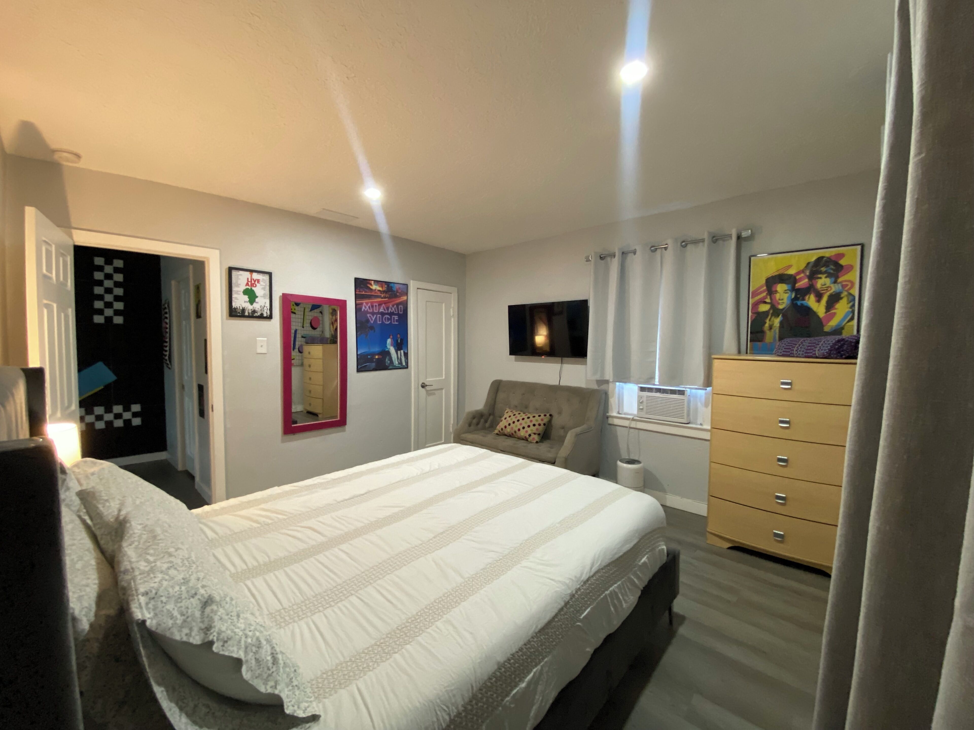 Un dormitorio con cama, cómoda, televisor y carteles en la pared.