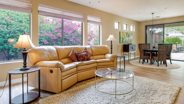 A living room with a beige sofa, a glass coffee table, and a white shag rug. There are two lamps, a dining table with chairs, and a view of a garden with pink flowers through large windows. - Snowbirds Welcome! Anthem Gem w/ Fairway Views (Anthem)