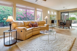 A living room with a beige sofa, a glass coffee table, and a white shag rug. There are two lamps, a dining table with chairs, and a view of a garden with pink flowers through large windows. - Snowbirds Welcome! Anthem Gem w/ Fairway Views (Anthem)