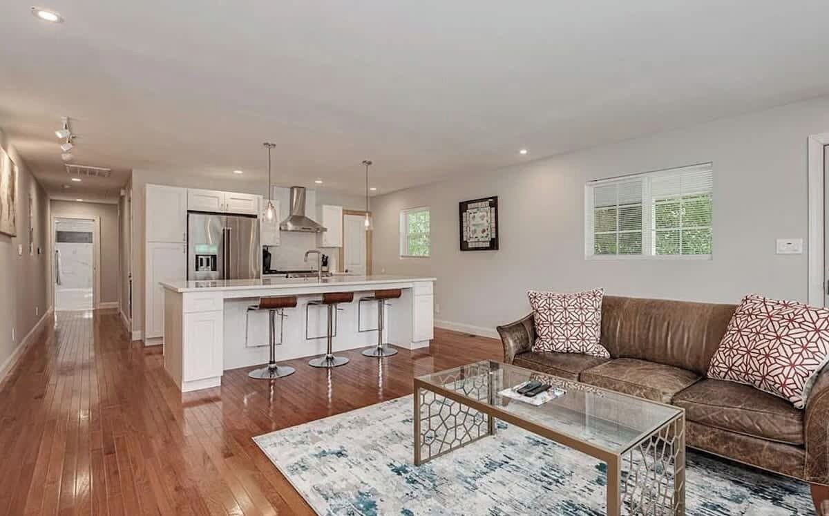 A modern kitchen with white cabinets, a central island with stools, a glass coffee table, and a brown leather sofa with patterned cushions.
