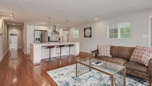 A modern kitchen with white cabinets, a central island with stools, a glass coffee table, and a brown leather sofa with patterned cushions. - Abbott Park 5 minutes to South End- 2 BR (Charlotte)