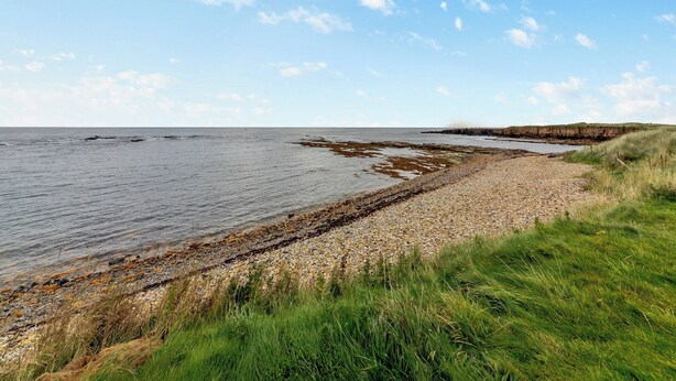 A coastal landscape with a pebbly beach, grassy dunes, and a clear sky. - Longbeach meets all your needs for a coastal family retreat. (Beadnell)