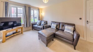 A living room with a large flat-screen TV on a wooden stand, a leather sectional sofa with cushions, and a floor lamp. - Longbeach meets all your needs for a coastal family retreat. (Beadnell)