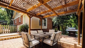An outdoor patio with a wooden pergola, a brick barbecue grill, and a dining table with chairs. - Comfy cozy laid back cottage (Charlotte)