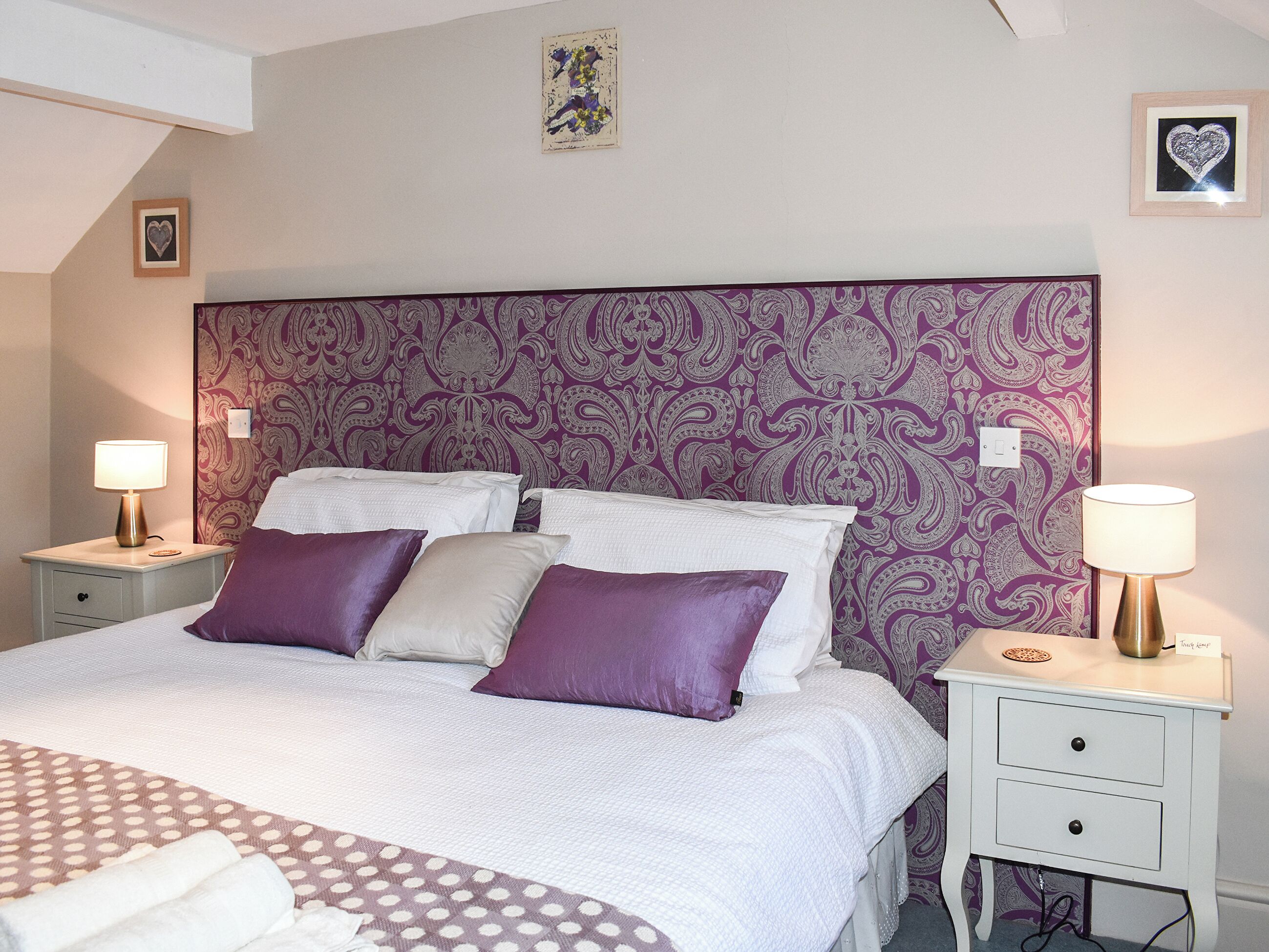 Cama bem arrumada com cabeceira estampada, duas mesas de cabeceira com abajures e uma parede com decoração em formato de coração.