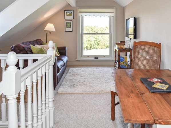 Un salon chaleureux avec un canapé en cuir marron, un escalier blanc, une table à manger en bois et une grande fenêtre donnant sur les arbres.