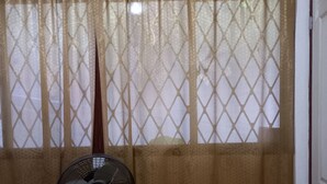Um quarto com um ventilador de coluna, uma janela com cortinas estampadas e uma porta.