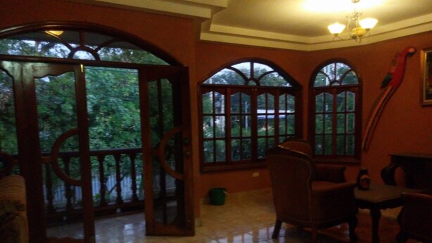 Um quarto com uma grande porta arqueada que dá para uma varanda, uma cadeira de madeira, uma mesa pequena e uma janela grande com vista para árvores.