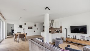 Un salon moderne avec un grand canapé, un coin repas comprenant une table et des chaises, une cuisine avec des meubles blancs et un téléviseur à écran plat fixé au mur.
