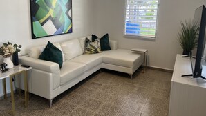 A modern living room with a white sectional sofa, a coffee table, a flat-screen TV, a potted plant, and a large abstract painting. - Juana Diaz Spacious Cozy Home Mango trees and avocado. (San Juan)