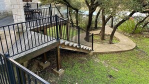 Une maison en pierre avec une terrasse en bois, un escalier en métal noir et un arbre en arrière-plan.
