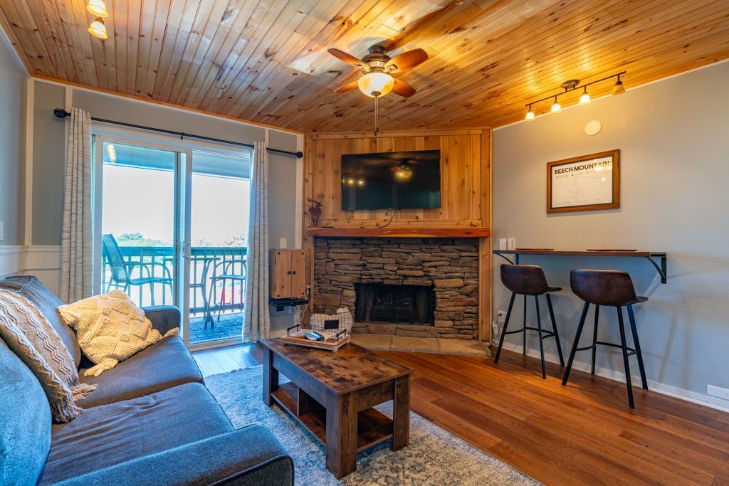 A cozy living room with a stone fireplace, a ceiling fan, and a bar area with stools.