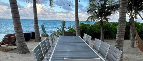 A beachside dining area with a long table and chairs, surrounded by palm trees and overlooking the ocean.