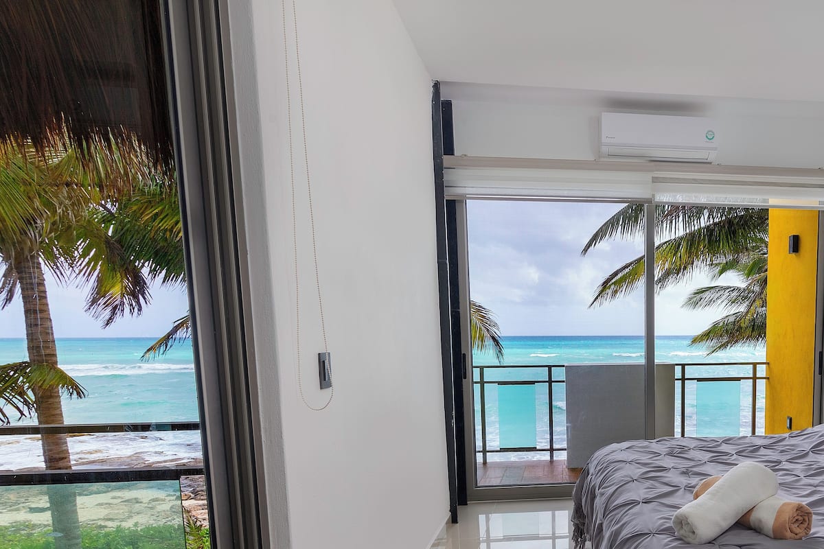 Ein Schlafzimmer mit großem Fenster mit Blick auf einen Strand, einem Bett mit einem zusammengerollten Handtuch und einem Balkon mit Meerblick.