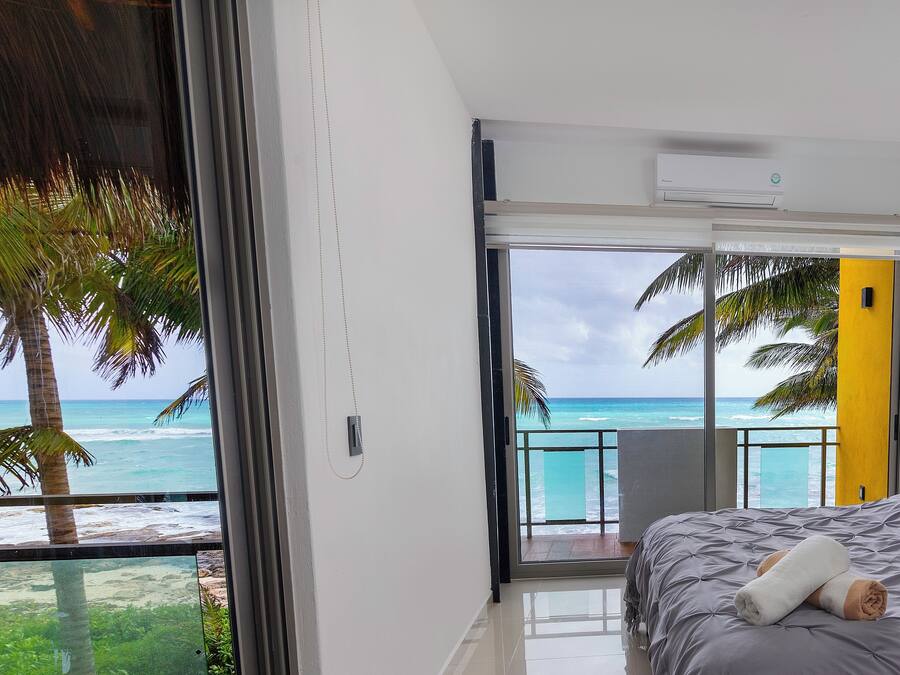 A bedroom with a large window overlooking a beach, a bed with a rolled towel, and a balcony with a view of the ocean.