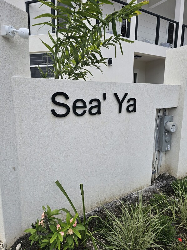 Una señal con el nombre 'Sea'Ya' sobre una pared con textura, rodeada de follaje y un edificio moderno al fondo.