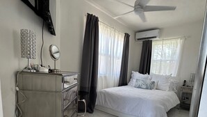 A hotel room with a bed, a ceiling fan, an air conditioning unit, a dresser, a mirror, and a window with curtains. - Brand New- Modern Sea' Ya Villa (Fitts Village)
