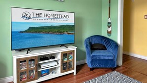 A living room with a television displaying 'The Homestead Oceanfront Rooms & Suites', a blue chair, a wooden cabinet with shelves, and a wall-mounted guitar.