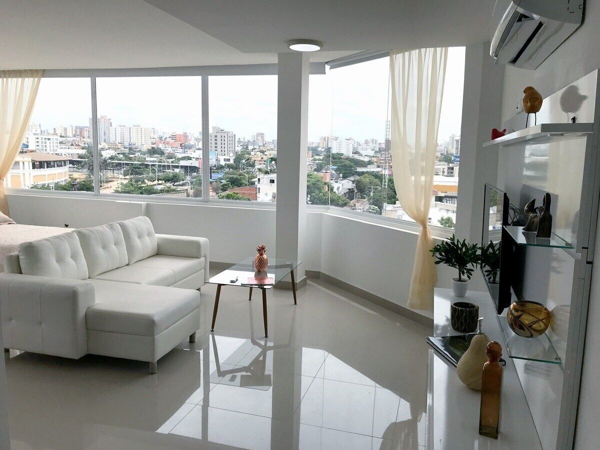 A modern living room with a white sofa, a glass coffee table, and a city view through large windows.