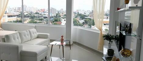 A modern living room with a white sofa, a glass coffee table, and a city view through large windows.
