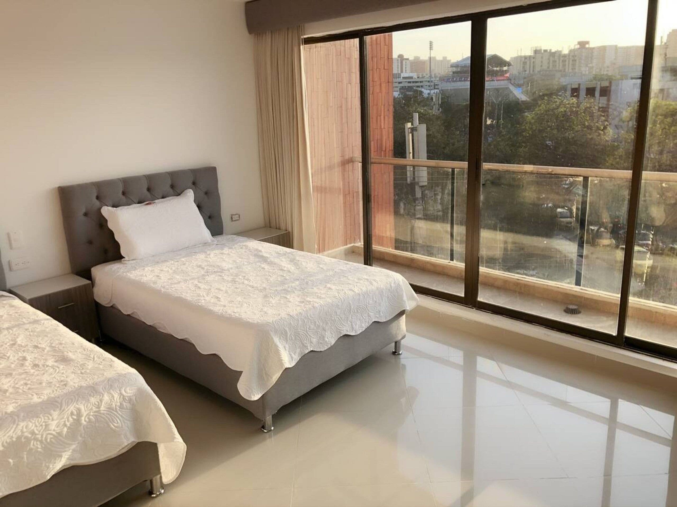 A bedroom with two beds, a large window with a view of buildings, and a balcony.