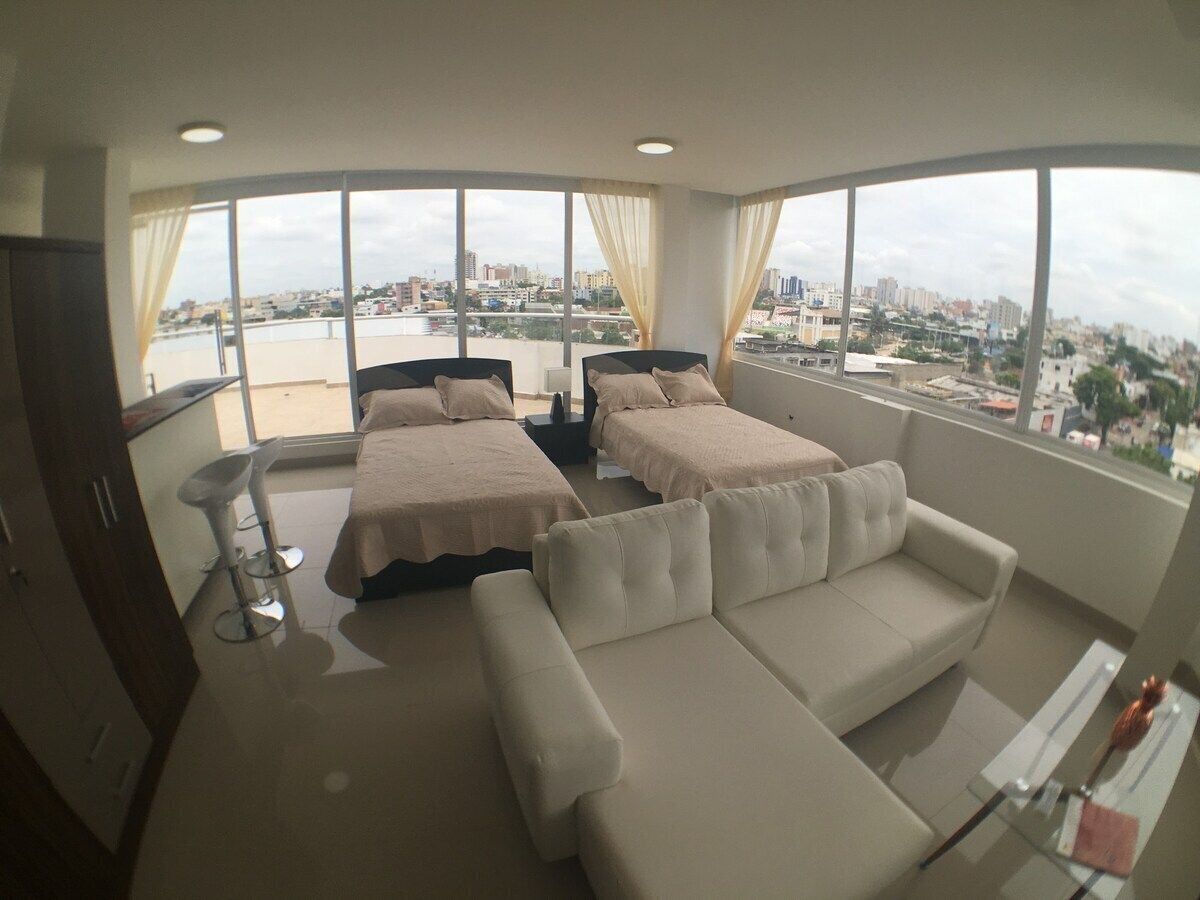 Habitación de hotel con dos camas, un sofá, un taburete de bar y ventanas amplias con vistas a la ciudad.
