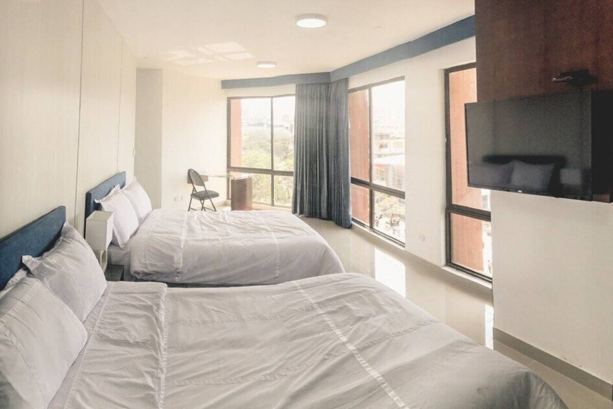 A hotel room with two beds, a TV, and a view of the city through large windows.