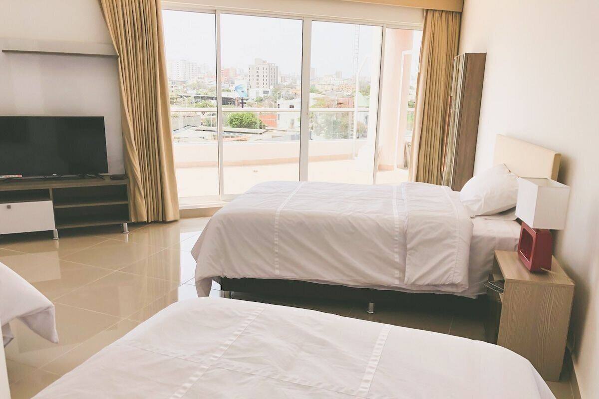 A hotel room with two beds, a TV, a desk, and a view of the city through large windows.