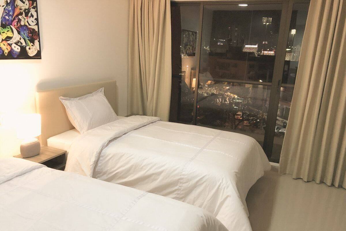A hotel room with two beds, a nightstand, a lamp, and a large window with curtains.