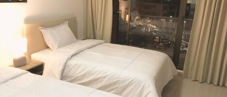 A hotel room with two beds, a nightstand, a lamp, and a large window with curtains.