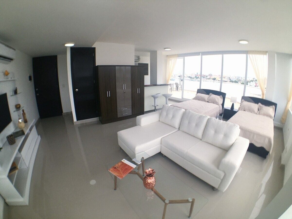 A modern living room with a white sofa, a small coffee table, and a TV on a stand. There is a kitchen area with dark cabinets and a window with curtains.