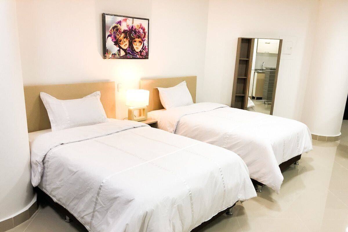 A hotel room with two single beds, a nightstand with a lamp, a painting on the wall, and a bathroom visible through an open door.