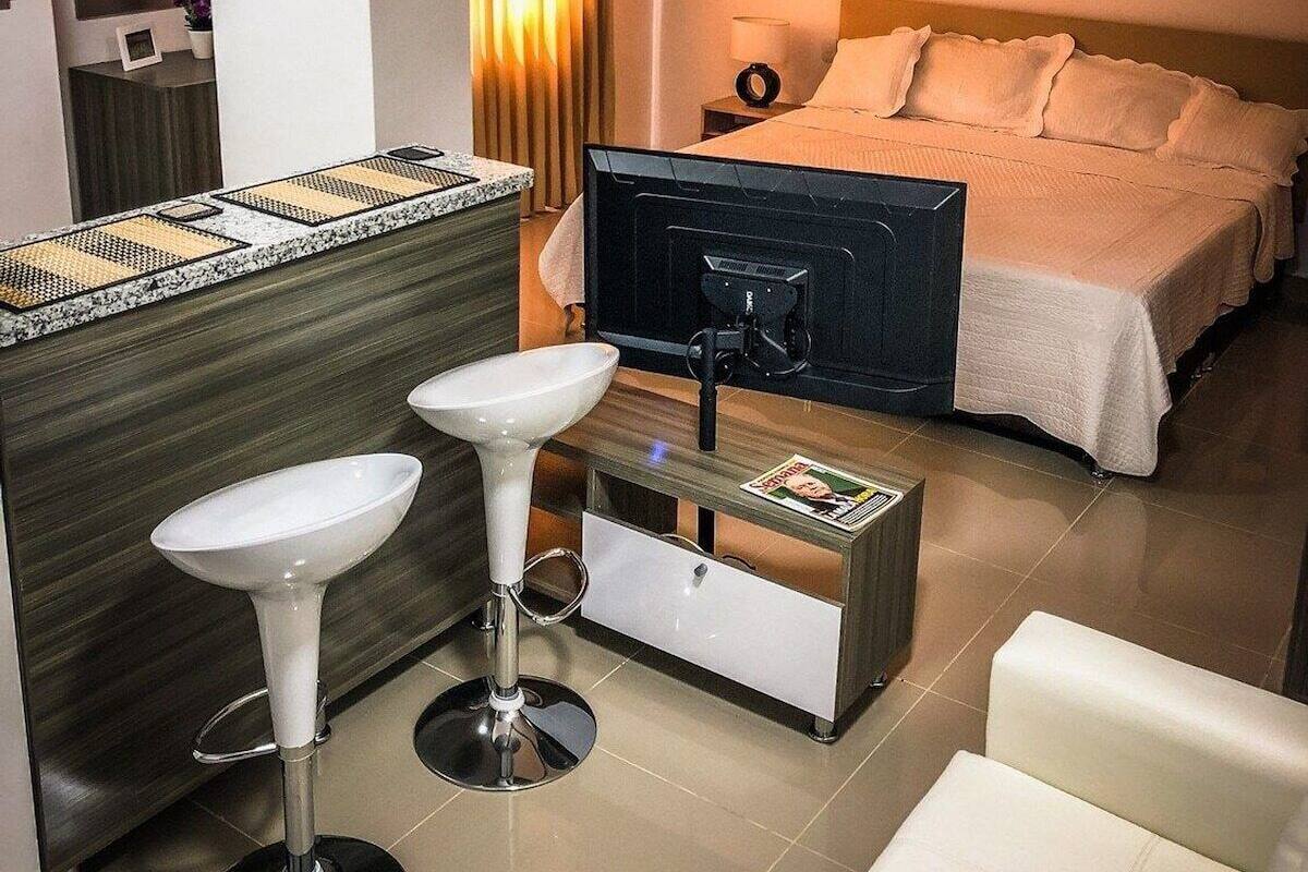 A hotel room with a bed, bedside table, lamp, and a small seating area with stools.