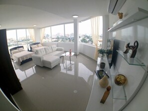 Amplia sala de estar con sofá blanco, una mesa de centro y un televisor sobre un mueble bajo. Amplias ventanas ofrecen vistas a la ciudad.