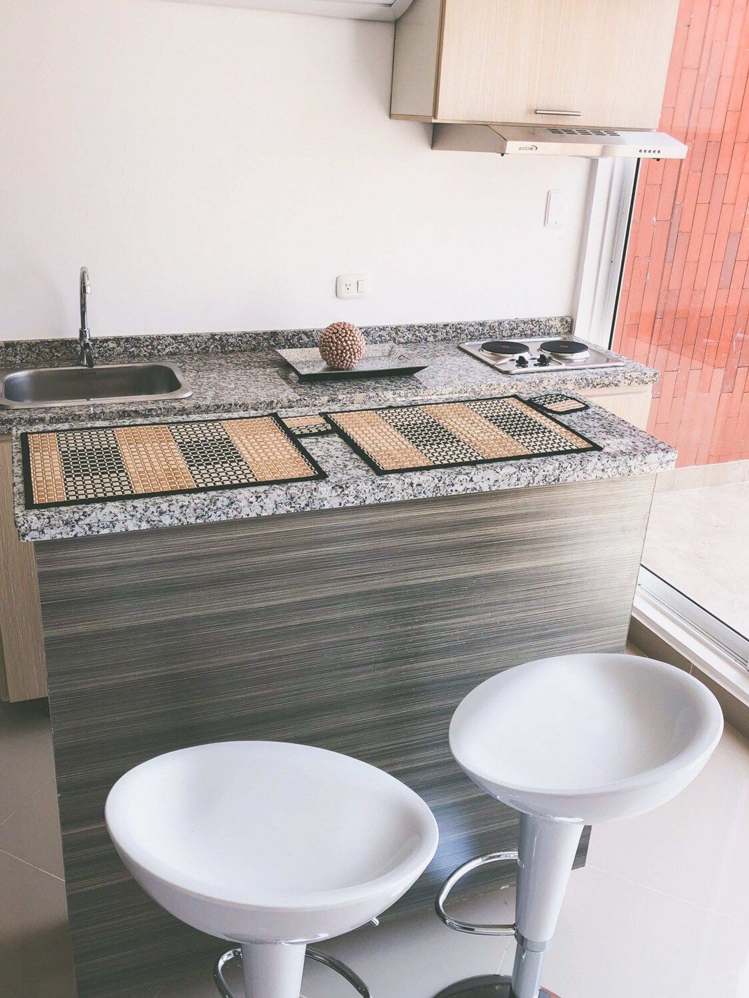 Cocina compacta con encimera de granito, estufa integrada, pileta y dos taburetes blancos.
