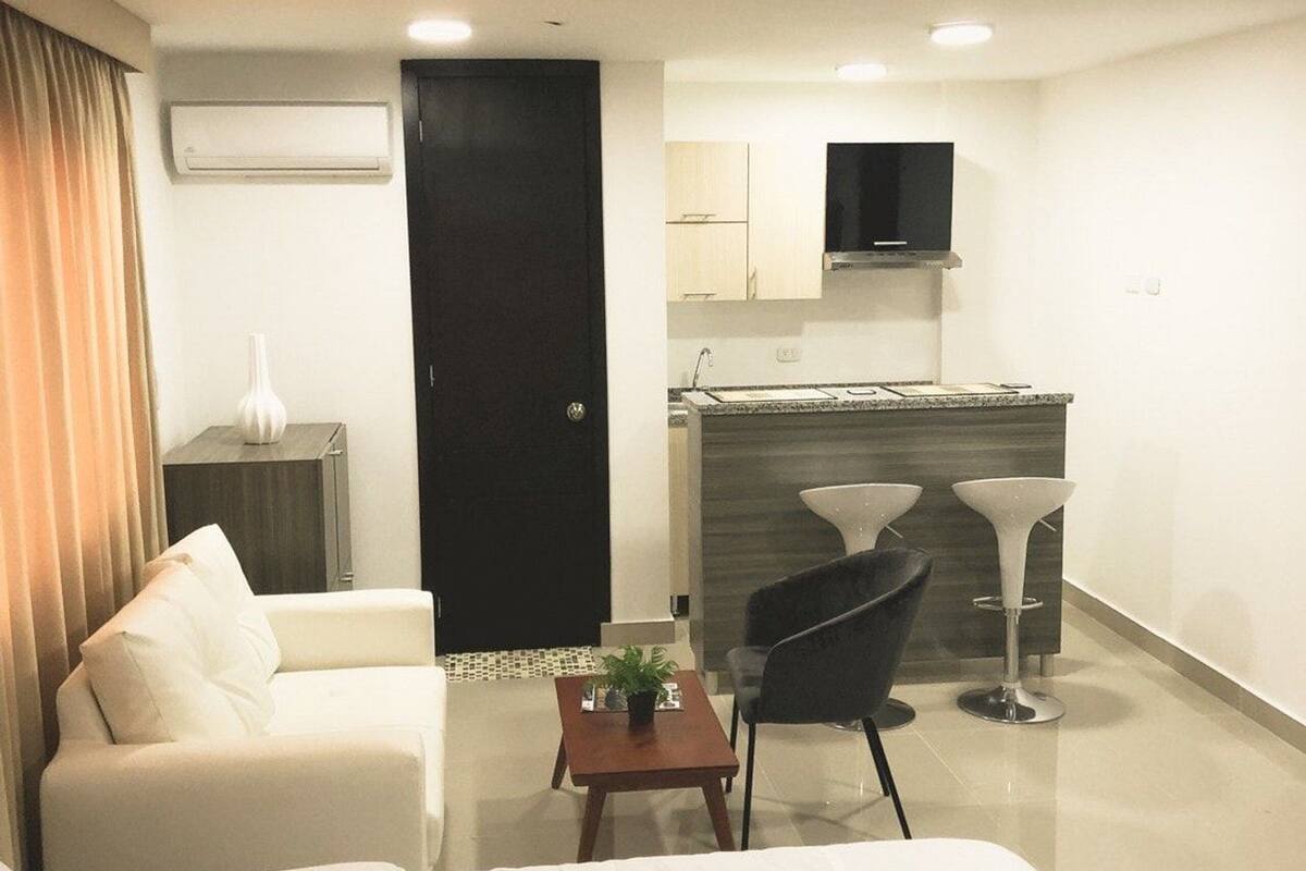 A modern hotel room with a bed, a sofa, a small table with a plant, a chair, and a kitchenette in the background.