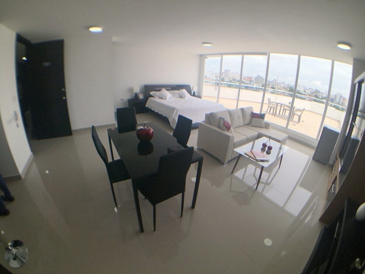 A modern hotel room with a bed, a dining table with chairs, a sofa, and a small coffee table.