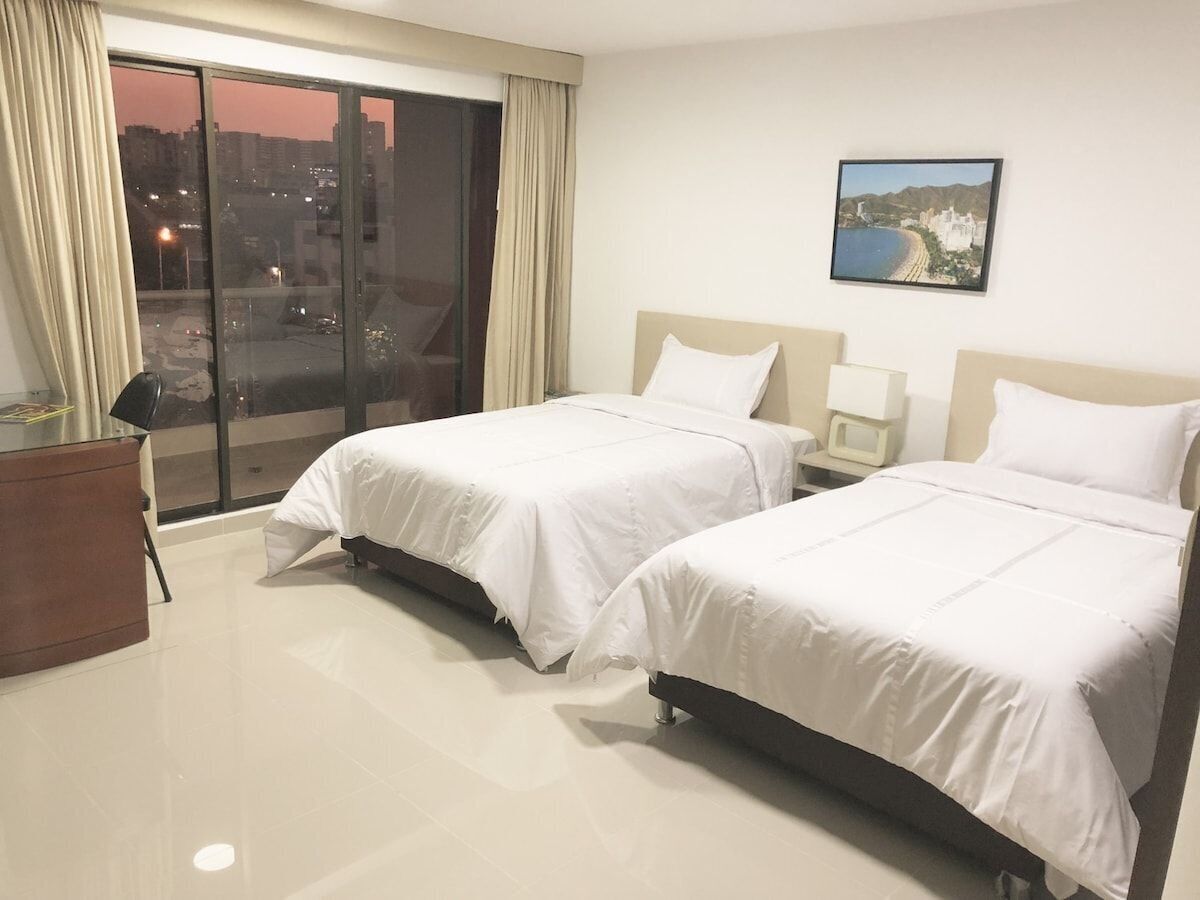 A hotel room with two beds, a large window with curtains, a desk, and a painting on the wall.